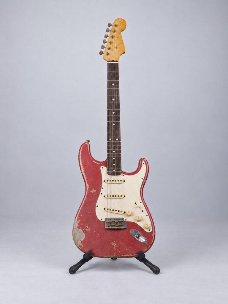 Red electric guitar on a stand with a worn finish, chipped down to bare wood along the edges.